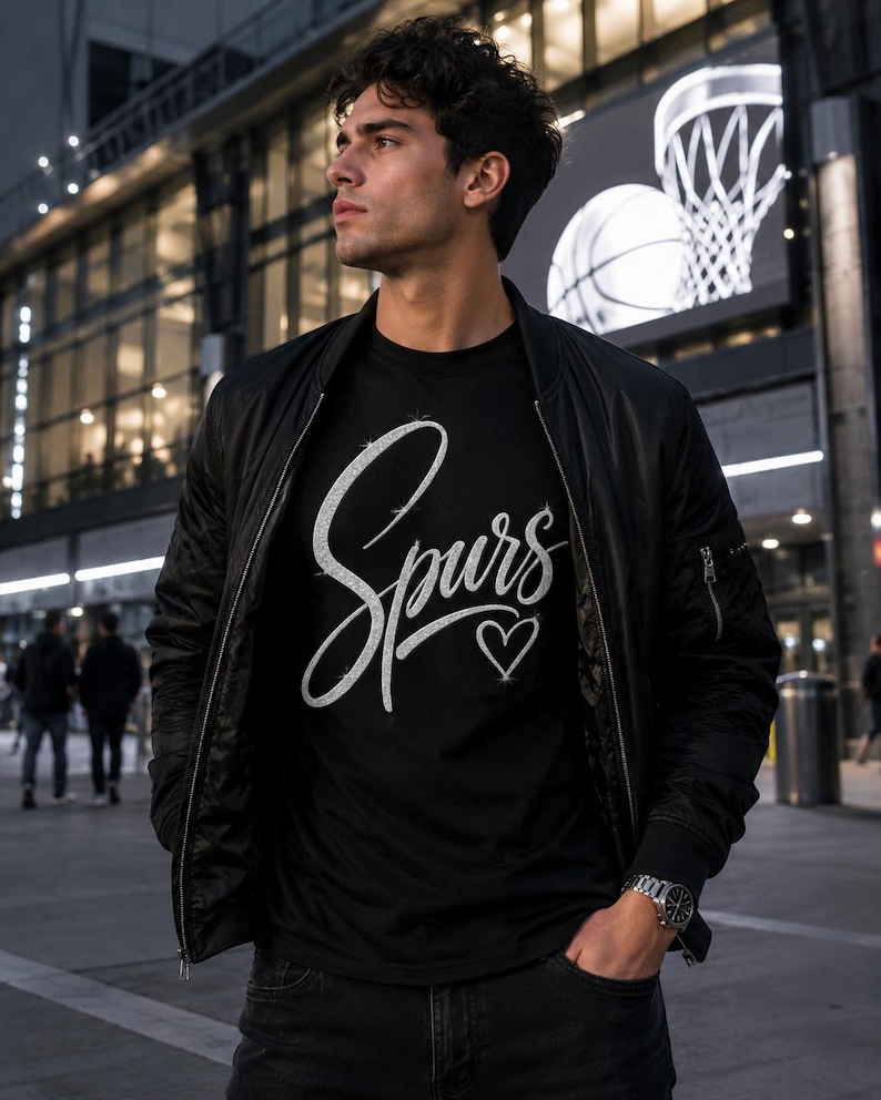 May include: A black t-shirt with the word "Spurs" in a cursive font and a heart symbol. The person is wearing a black bomber jacket and dark jeans. A basketball hoop and ball are visible in the background.