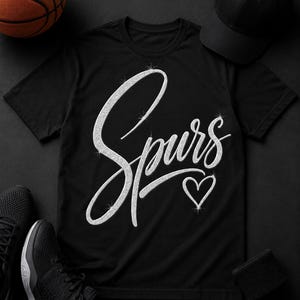 May include: Black t-shirt with the word "Spurs" in a sparkly, cursive font and a sparkly heart. A black baseball cap, black sneakers, a black wristband, and a basketball are arranged around the shirt on a black surface.