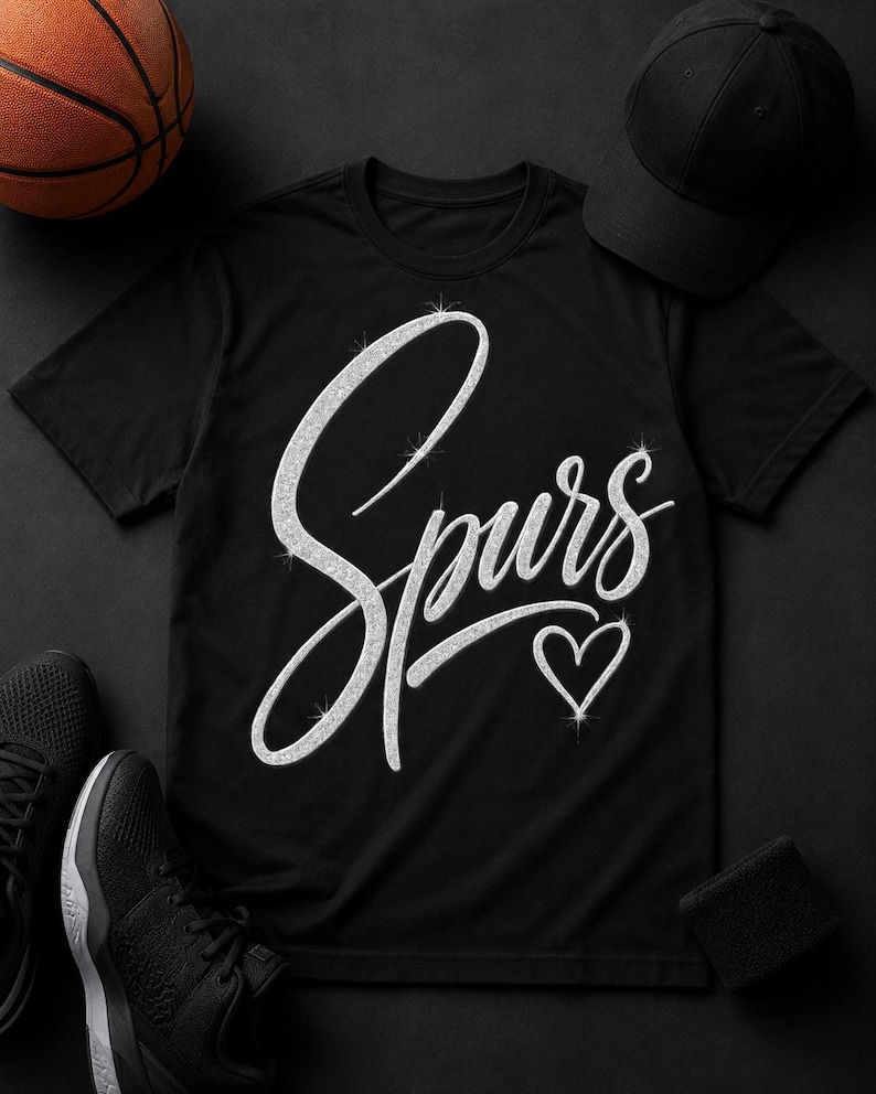 May include: Black t-shirt with the word "Spurs" in a sparkly, cursive font and a sparkly heart. A black baseball cap, black sneakers, a black wristband, and a basketball are arranged around the shirt on a black surface.