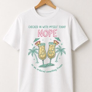 Può includere: Una t-shirt bianca a maniche corte con una grafica di due cocktail tropicali con cubetti di ghiaccio, fette di ananas e ombrellini. Il testo recita: CHECKED IN WITH MYSELF TODAY NOPE so we ordered something frozen, con illustrazioni di palme.