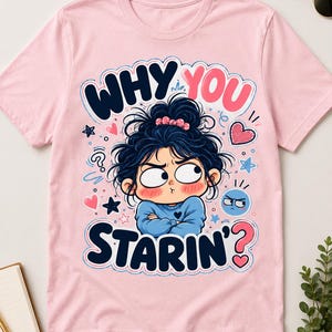 May include: A light pink t-shirt featuring a graphic illustration of a person with messy dark hair, a blue sweater, and a grumpy expression. The text "WHY YOU STARIN'?" is printed in bold dark blue and pink letters, surrounded by small hearts and stars.