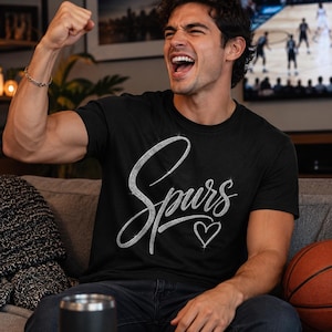 May include: Black t-shirt with the word "Spurs" in a silver, cursive font and a small heart. A person is wearing the shirt and cheering, with a basketball and a bowl of snacks nearby.