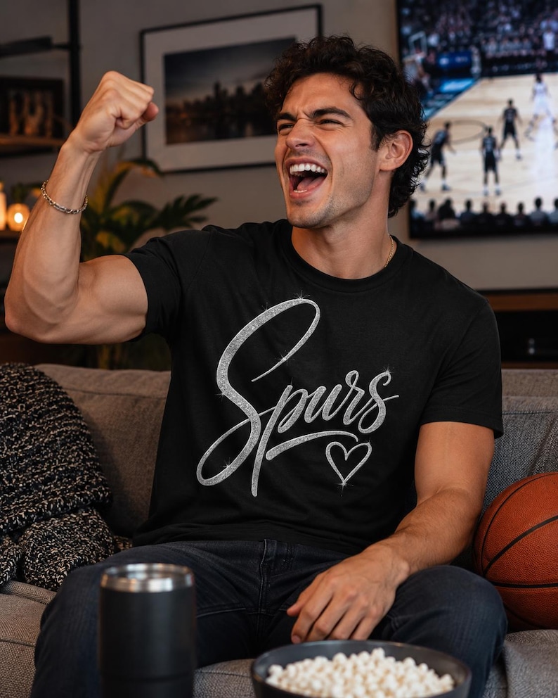 May include: Black t-shirt with the word "Spurs" in a silver, cursive font and a small heart. A person is wearing the shirt and cheering, with a basketball and a bowl of snacks nearby.