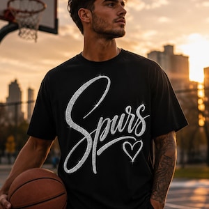 May include: Black t-shirt with the word "Spurs" in a sparkly silver script and a silver heart. The shirt is short-sleeved. A basketball and a basketball hoop are in the background.