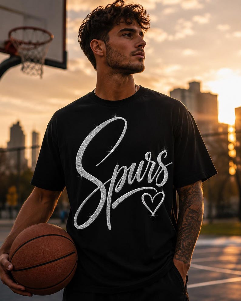 May include: Black t-shirt with the word "Spurs" in a sparkly silver script and a silver heart. The shirt is short-sleeved. A basketball and a basketball hoop are in the background.