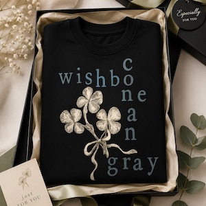 Könnte beinhalten: Schwarzes Sweatshirt mit dem blauen Schriftzug "wishbone can gray" und einer detaillierten Illustration eines Kleeblatt-Bouquets, das mit einer Schleife gebunden ist. Das Sweatshirt wird in einer Geschenkbox mit einer Karte "just for you" präsentiert.