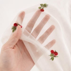 Puede incluir: Tela blanca transparente con cerezas rojas y hojas verdes bordadas. La delicada tela se sostiene, mostrando la transparencia y el bordado detallado de la fruta y el follaje.