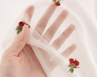 Puede incluir: Tela blanca transparente con cerezas rojas y hojas verdes bordadas. La delicada tela se sostiene, mostrando la transparencia y el bordado detallado de la fruta y el follaje.