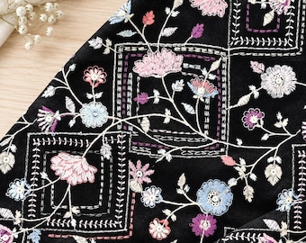 Op de afbeelding: Een zwarte stof met een complex geborduurd patroon van vierkante kaders en delicate bloemmotieven. Het borduurwerk gebruikt draden in tinten roze, lichtblauw, paars en wit, wat zorgt voor een gedetailleerd botanisch ontwerp op het oppervlak.