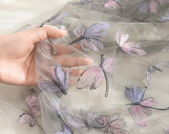 Op de afbeelding: Een transparante, lichtgrijze tule stof met een geborduurd patroon van libellen. De libellen zijn gedetailleerd met glinsterende pailletten in tinten zachtroze, lavendel en zilver, wat zorgt voor een delicaat, getextureerd oppervlak.