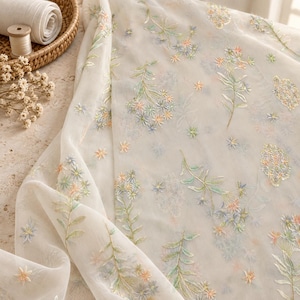 Könnte beinhalten: Ein transparenter, cremeweißer Stoff mit zarter Blumenstickerei in sanften Pastelltönen wie Pfirsich, Hellblau und Salbeigrün. Das Muster besteht aus verstreuten botanischen Zweigen und kleinen sternförmigen Blüten auf dem durchscheinenden Material.