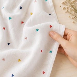 Puede incluir: Tela blanca con corazones bordados en varios colores, incluyendo rojo, azul, verde y amarillo. La tela es sostenida, mostrando su textura y el patrón de corazones dispersos. La tela parece ligera y adecuada para diversos usos.