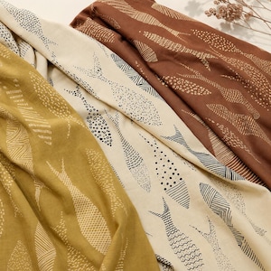 Pode incluir: Três peças de tecido dobradas com um padrão repetitivo de peixes. Uma peça é amarelo-mostarda, uma é creme com desenhos de peixes em tinta preta e uma é castanho-escura com desenhos de peixes em tinta branca. Cada peixe tem uma textura.