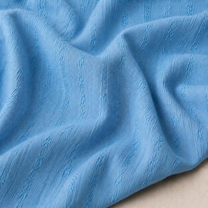 Peut inclure: Un tissu bleu clair présentant un motif texturé vertical composé de colonnes tricotées répétitives en forme de câbles. Le matériau a un aspect souple et drapé avec des plis doux qui soulignent le design tissé cohérent sur la surface.
