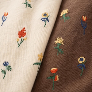 Könnte beinhalten: Nahaufnahme von zwei Stoffmustern, eines cremefarben und eines braun, beide mit farbenfrohen Blumenmustern bestickt. Die Blumen umfassen rote, gelbe, blaue und orangefarbene Blüten mit grünen Stielen und Blättern. Der Stoff hat ein strukturiertes Aussehen.