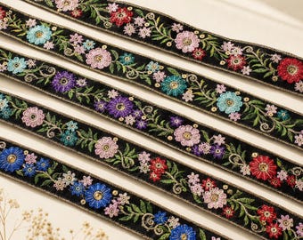 Op de afbeelding: Vier stroken zwarte stoffen bies met ingewikkeld bloemenborduursel in tinten rood, roze, blauw, paars en turkoois. Elke strook is versierd met groene bladeren, gouden metallic draadaccenten en kleine lovertjes in het midden van de bloemen.