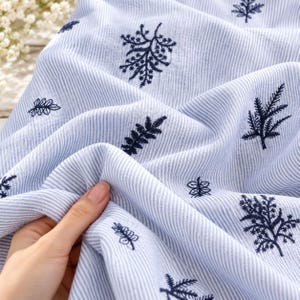 Könnte beinhalten: Ein hellblau-weiß gestreifter Stoff mit verstreuten, dunkelblau gestickten botanischen Motiven. Das Material hat eine weiche, strukturierte Optik mit Falten, die das sich wiederholende Muster aus verschiedenen kleinen Blatt- und Zweigdesigns betonen.