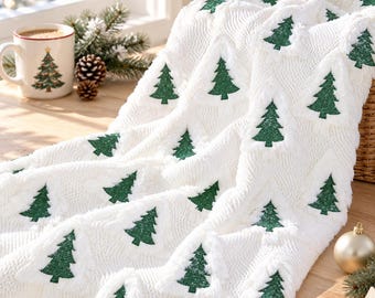 Op de afbeelding: Een witte gebreide plaid met een herhalend patroon van donkergroene kerstbomen. De stof heeft een gestructureerd kabelgebreid uiterlijk waarbij de bomen in het weefsel zijn verwerkt. De deken is gedrapeerd in zachte plooien.