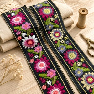 Könnte beinhalten: Zwei schwarze Stoffbänder mit lebendiger Blumenstickerei in Magenta, Pink, Gelb, Weiß und Blau, akzentuiert durch grüne Blätter und feine goldene Metallic-Linien, die entlang der Kanten des dekorativen Bandes verlaufen.