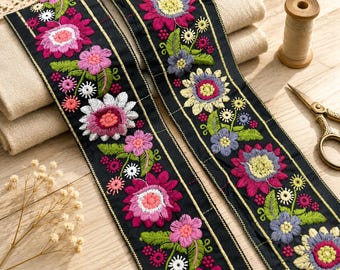 Op de afbeelding: Twee stroken zwarte stoffen bies met levendig bloemenborduursel in tinten magenta, roze, geel, wit en blauw, geaccentueerd met groene bladeren en dunne gouden metallic lijnen langs de randen van het decoratieve lint.