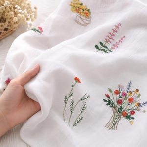 Puede incluir: Tela blanca con bordados florales coloridos. El bordado incluye un ramo de flores, una sola flor roja y otros diseños florales. La tela está siendo sostenida por una mano.
