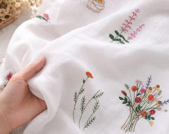 Puede incluir: Tela blanca con bordados florales coloridos. El bordado incluye un ramo de flores, una sola flor roja y otros diseños florales. La tela está siendo sostenida por una mano.