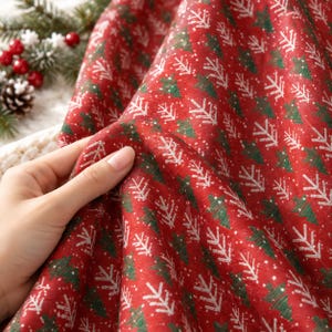 Könnte beinhalten: Ein roter Stoff mit einem sich wiederholenden Muster aus stilisierten grünen und weißen Weihnachtsbäumen. Das Material hat eine texturierte Oberfläche mit kleinen weißen Punkten, die über das Design verteilt sind.