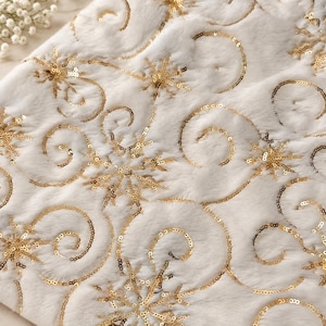 Könnte beinhalten: Ein weißer Webpelzstoff mit einem komplizierten Muster aus goldenen Pailletten-Schneeflocken und geschwungenen Linien. Die weiche, flauschige Textur des weißen Materials bildet einen Kontrast zum metallischen Glanz der aufgestickten Pailletten.