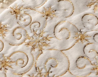Op de afbeelding: Een witte imitatiebontstof met een ingewikkeld patroon van gouden pailletten sneeuwvlokken en wervelende lijnen. De zachte, pluizige textuur van het witte materiaal contrasteert met de metallic glans van de pailletten op het oppervlak.