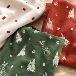 Könnte beinhalten: Nahaufnahme von gefaltetem Stoff mit Weihnachtsmotiven in Creme, Grün und Rost. Der cremefarbene Stoff hat rote Sockenstickereien. Der grüne Stoff zeigt weiße, gestickte Hirsche, Schneeflocken und Weihnachtsbäume. Der rostfarbene Stoff hat weiße Weihnachtsbaum- und Schneeflockenstickereien.
