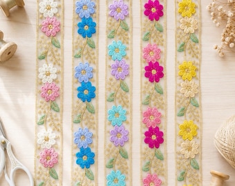 Op de afbeelding: Vijf verticale stroken van transparant kanten lint met geborduurde bloemmotieven in diverse kleuren, waaronder wit, roze, blauw, paars en geel. Elke strook is voorzien van een gouden metallic draadrand en groene geborduurde blaadjes.