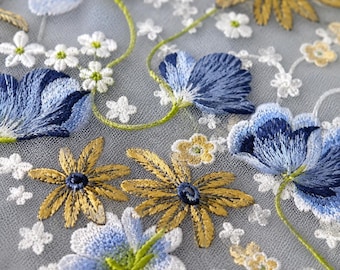 Op de afbeelding: Een transparante grijze mesh-stof met ingewikkeld borduurwerk van blauwe en witte bloemen, goudgele madeliefachtige bloesems en dunne groene stengels. Het bloemmotief is dicht verspreid over het doorschijnende materiaal met gedetailleerd stikwerk.