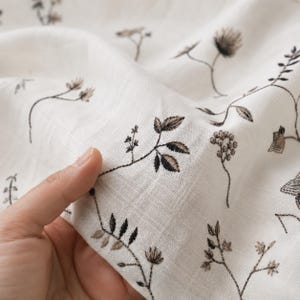 Puede incluir: Una pieza de tela color blanco roto con un delicado patrón de bordado repetitivo de elementos botánicos en marrón oscuro, incluyendo pequeñas hojas, tallos y racimos florales, distribuidos por la superficie texturizada del material ligero.