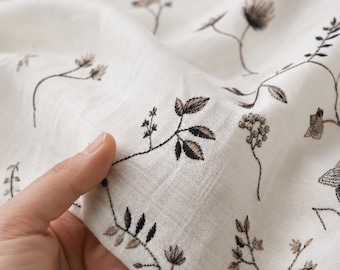 Puede incluir: Una pieza de tela color blanco roto con un delicado patrón de bordado repetitivo de elementos botánicos en marrón oscuro, incluyendo pequeñas hojas, tallos y racimos florales, distribuidos por la superficie texturizada del material ligero.