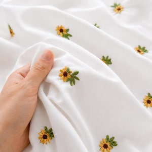 Peut inclure: Tissu blanc avec des broderies de tournesols jaunes et bruns et de feuilles vertes. Le tissu est doux et fluide, une main tenant une partie du matériau. Les tournesols sont espacés uniformément sur le tissu.