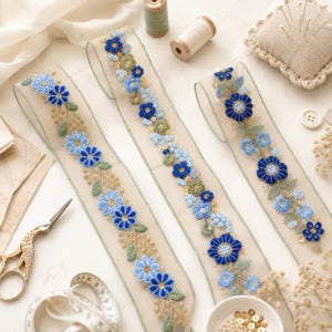 Könnte beinhalten: Drei Streifen aus transparentem, hellem Band mit aufwendigen Stickereien aus blauen Blumen, grünen Blättern und kleinen goldenen Pailletten. Das florale Muster ist als durchgehendes Rankendesign entlang der Länge jeder Stoffborte angeordnet.