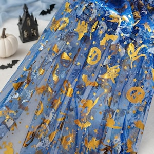Könnte beinhalten: Ein transparenter blauer Stoff mit einem metallischen Goldfoliendruck von Halloween-Motiven wie Kürbissen, Fledermäusen, Sternen und Hexenhüten. Das durchscheinende Material ist in vertikalen Falten drapiert und zeigt das Muster.