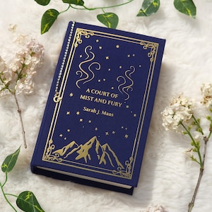 Op de afbeelding: Een marineblauw hardcover boek met de titel "A Court of Mist and Fury" van Sarah J. Maas. Het boek heeft gouden letters en illustraties, waaronder bergen en sterren. Een decoratieve zilveren bladwijzer is bevestigd.