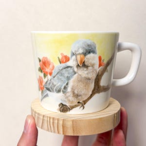 Puede incluir: Una taza de cerámica blanca con una ilustración en acuarela de un pequeño pájaro gris y beige posado en una rama marrón. El fondo incluye un suave tono amarillo y varias flores de color naranja brillante. La taza tiene un asa curva a la derecha.