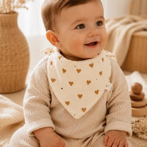 Peut inclure: Un bavoir triangulaire pour bébé en mousseline de couleur crème, orné d'un motif répétitif de petits cœurs jaune moutarde. Le bavoir se noue autour du cou et comporte une petite étiquette en tissu blanc sur le bord latéral.