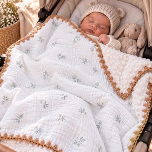 Peut inclure: Une couverture pour bébé en mousseline blanche avec un imprimé botanique vert, dotée d'un envers texturé couleur crème et d'une bordure crochetée beige. La couverture recouvre un nourrisson endormi portant un bonnet à oreilles d'ours.