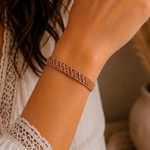 Op de afbeelding: Een beige geweven armband met een gedetailleerd, herhalend knooppatroon. De armband is gemaakt van dun, koordachtig materiaal en behoudt een consistente, smalle breedte rond de pols, wat een getextureerd en ingewikkeld ontwerp laat zien.