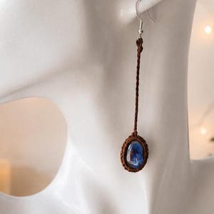 Puede incluir: Un pendiente largo con un cordón trenzado marrón sujeto a un gancho de tono plateado. En la base del cordón cuelga un pequeño colgante ovalado con un borde tejido de color marrón oscuro que rodea una piedra azul pulida con vetas blancas.