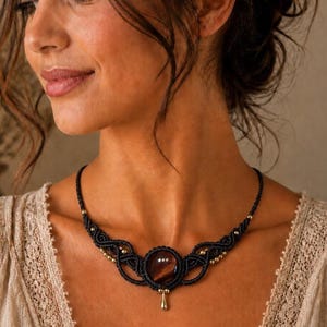 May include: A black macrame necklace featuring a central round dark brown gemstone, intricate knotted cord patterns, and small metallic gold-toned beads. A single gold-toned teardrop-shaped charm hangs from the bottom of the central stone setting.