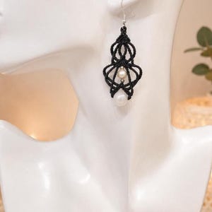 May include: A dangling earring featuring intricate black macrame cord knotted into a symmetrical, openwork geometric shape. The design incorporates a small, round white pearl at the center and a larger, round white pearl at the bottom tip.