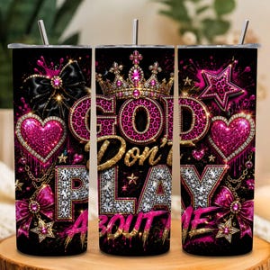 May include: A cylindrical tumbler with a black background featuring a pink leopard print, gold crown, and diamond-encrusted text that reads GOD DON'T PLAY ABOUT ME. The design includes pink hearts, black bows, and gold stars with metallic accents.