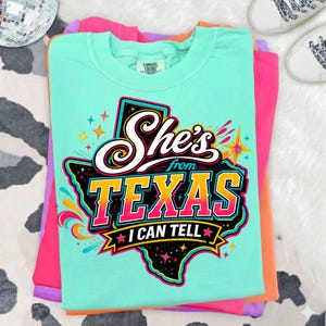 Pu&ograve; includere: Una t-shirt verde menta con una grafica dello stato del Texas in nero con accenti colorati. Il testo "She's from TEXAS I CAN TELL" &egrave; scritto in bianco, giallo e rosa, circondato da forme a stella e vortici astratti e colorati.