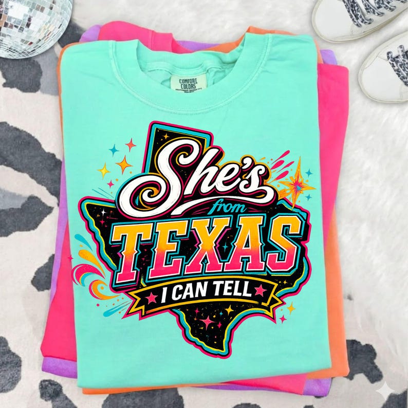 Pu&ograve; includere: Una t-shirt verde menta con una grafica dello stato del Texas in nero con accenti colorati. Il testo "She's from TEXAS I CAN TELL" &egrave; scritto in bianco, giallo e rosa, circondato da forme a stella e vortici astratti e colorati.