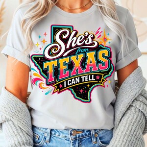 Pu&ograve; includere: Una t-shirt grigio chiaro a maniche corte con una grafica dello stato del Texas in nero con un bordo colorato. Il testo recita She's from TEXAS I CAN TELL in lettere bianche, gialle e rosa, accentuato da stelle colorate e forme astratte.