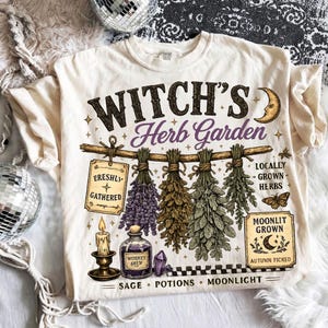 Op de afbeelding: Een crèmekleurig t-shirt met een grafische print met de tekst "WITCH'S Herb Garden". Het ontwerp bevat illustraties van hangende gedroogde kruiden, een kaars, een toverdrankflesje, een maansikkel en de teksten "FRESHLY GATHERED", "LOCALLY GROWN HERBS", "MOONLIT GROWN", "AUTUMN PICKED", "SAGE", "POTIONS" en "MOONLIGHT".
